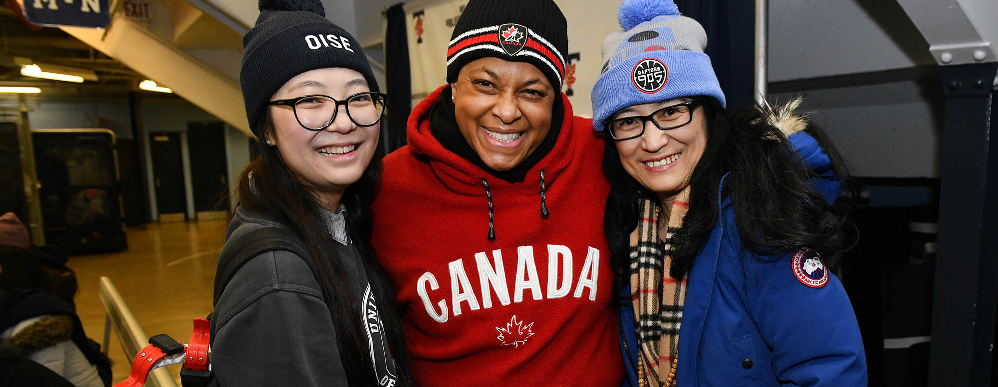 Three people smiling and close together. Each person is wearing winter clothing and a toque. 