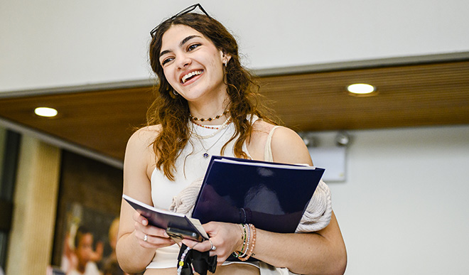 Woman with dark hair standing in the library, smiling as she holds a folder.