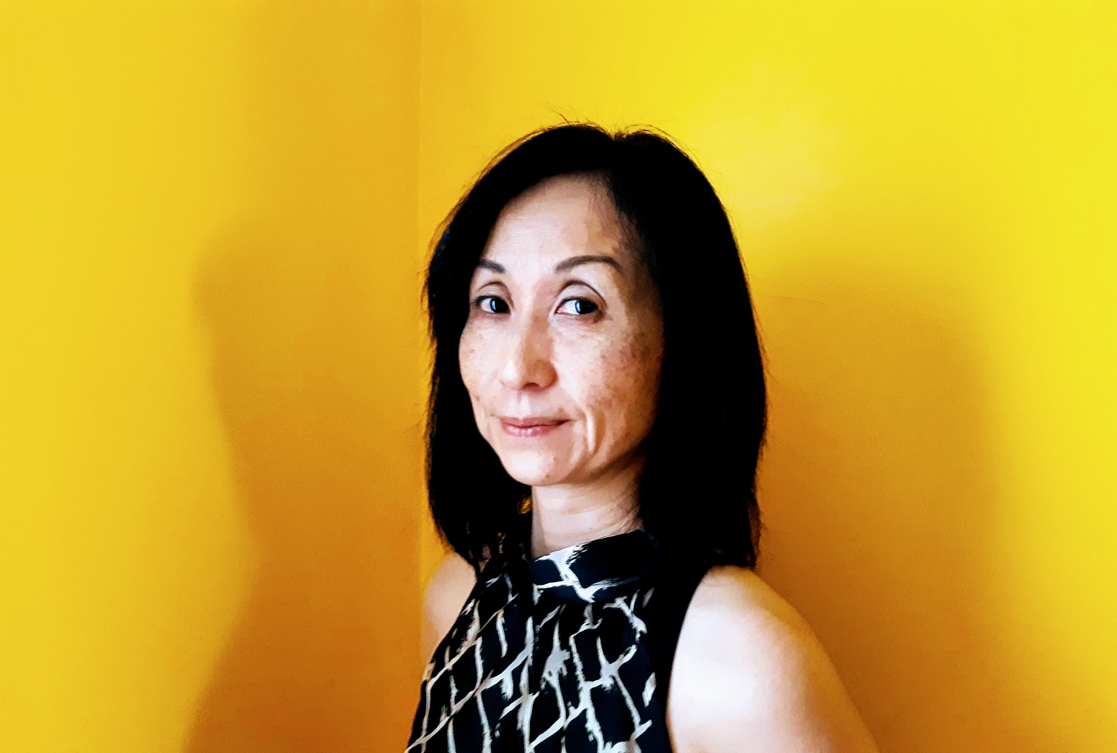 Woman against a yellow background, with dark shoulder length hair, and a black and white patterned shirt