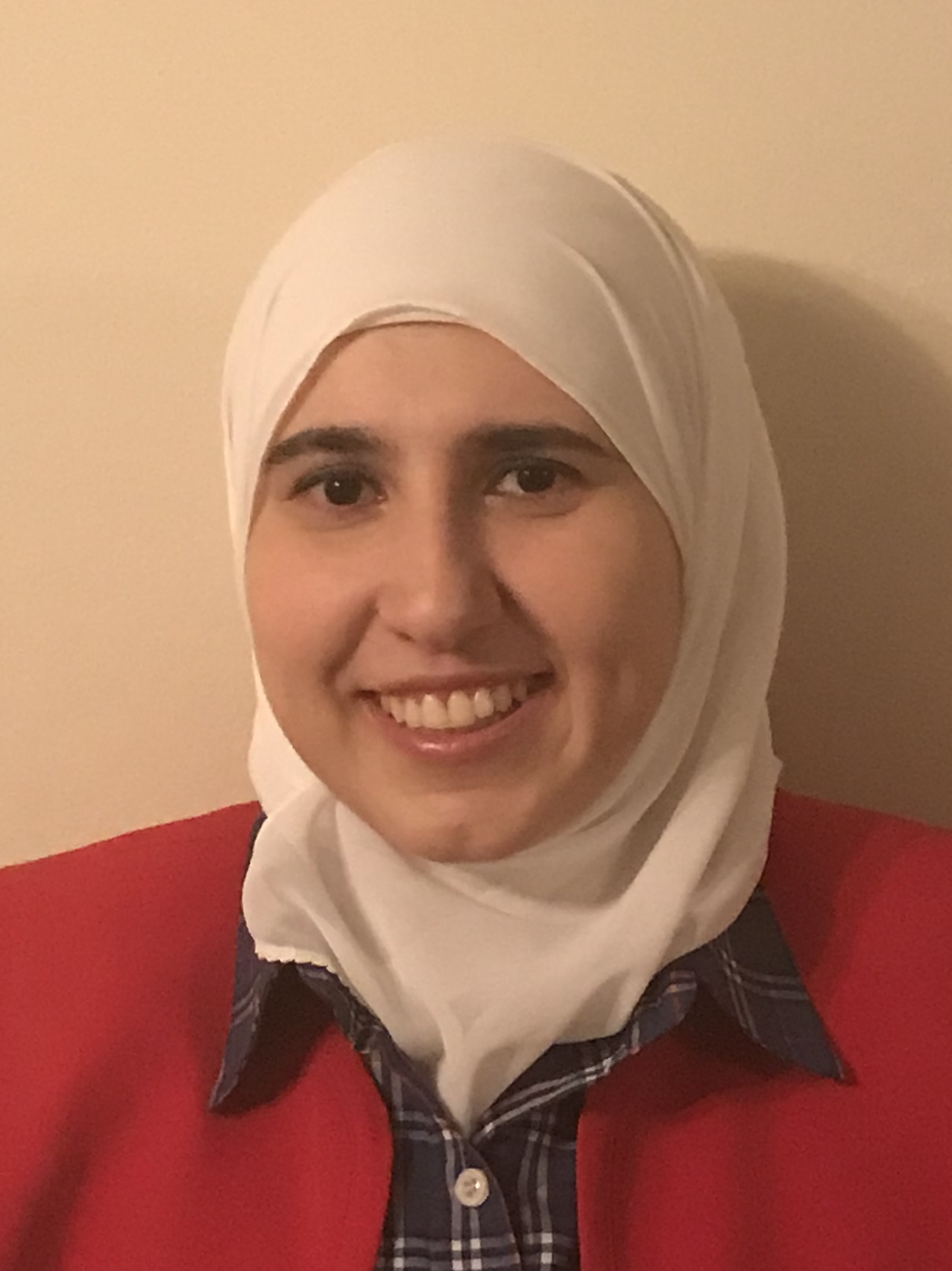 Woman in a white headscarf, smiling against a beige wall, wearing a red sweater