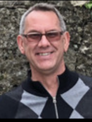 Man with short grey hair, smiling, wearing a grey diamond pattern zip-up sweater