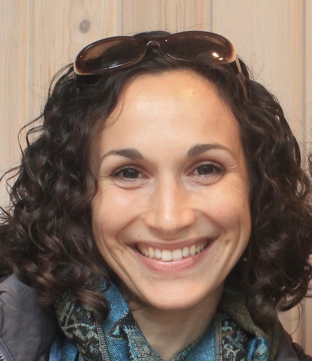 Smiling woman with curly brown, shoulder length hair, with sunglasses on her head