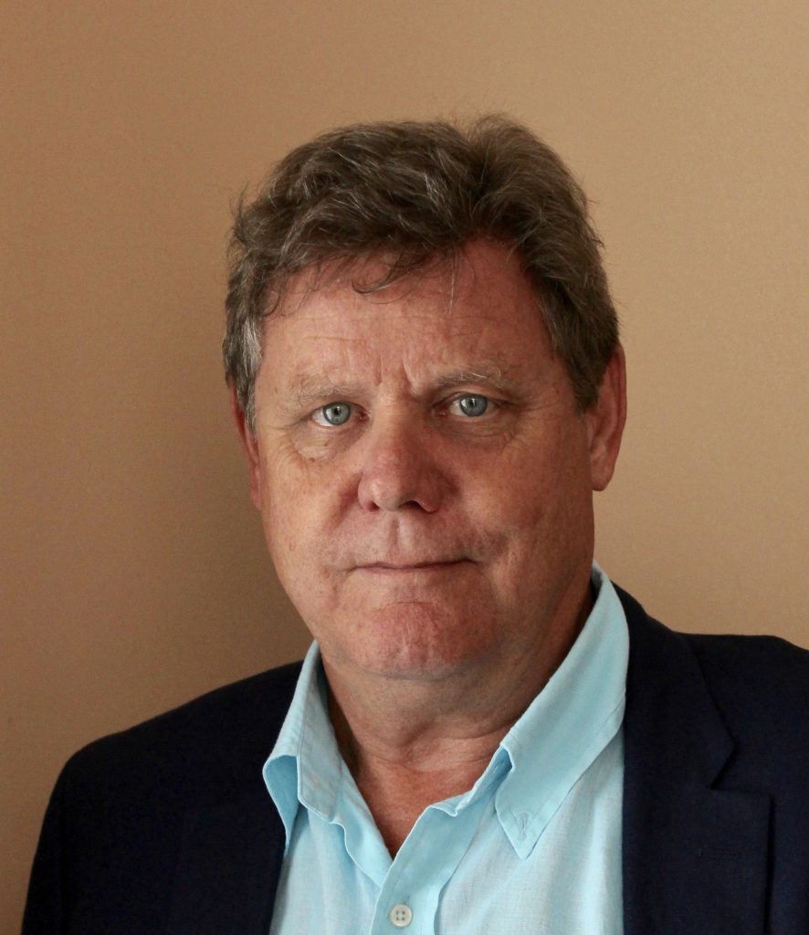 Short haired man against a beige background, wearing a black sportscoat and light blue dress shirt