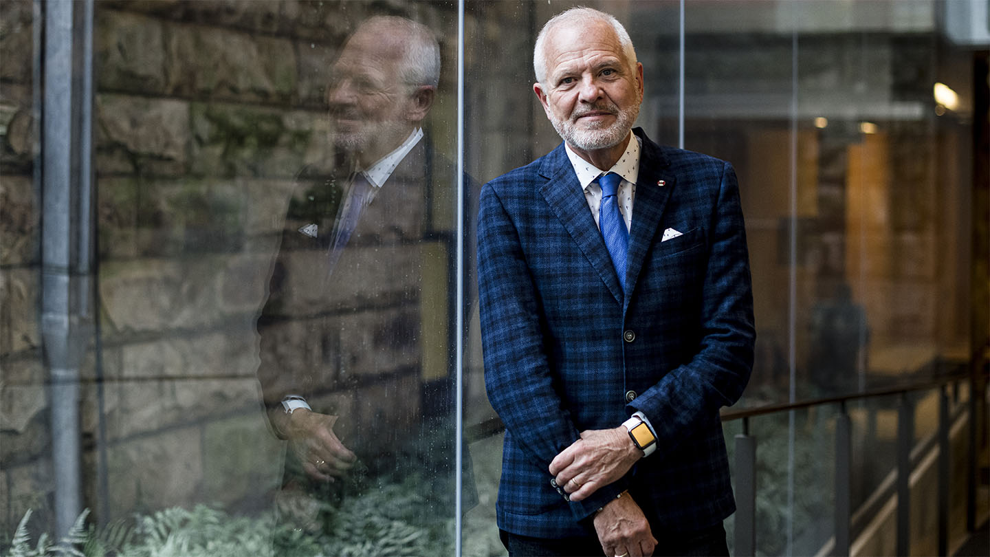 OISE Dean Normand in a blue suite smiling as he leans against a glass wall