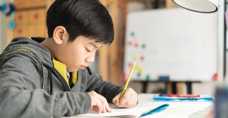 East Asian teenager is writing on a piece of paper with a pencil and looking thoughtful 