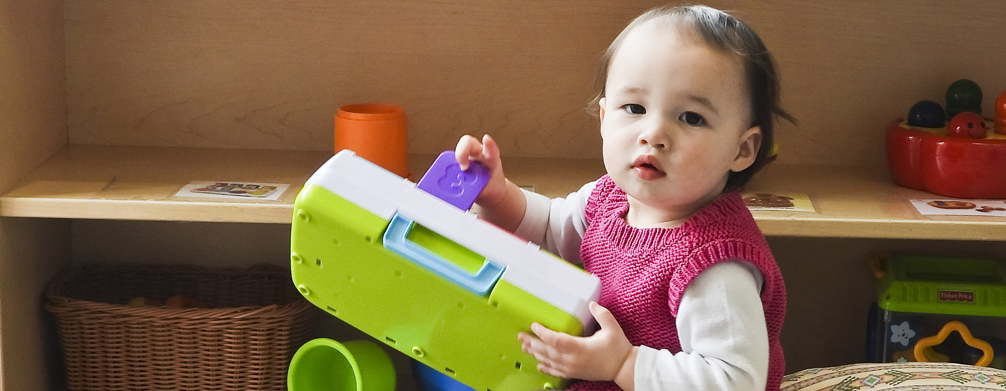 A child holding a green and white box, with the number 3 visible, in a daycare setting. 