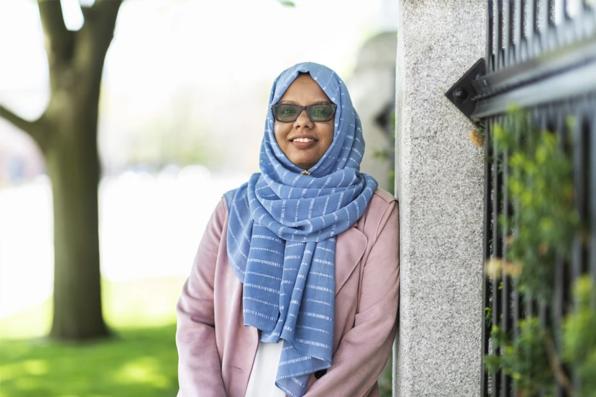 Mariam Abeid is wearing a blue hijab and sunglasses. She is smiling and is casually leaning against a pillar.