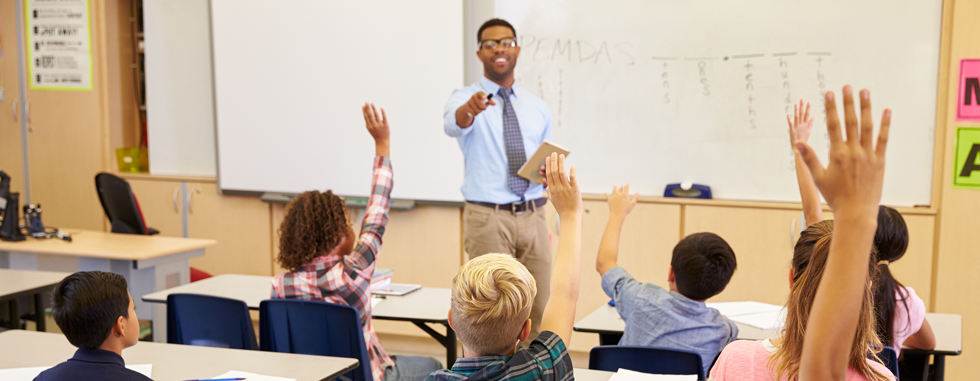 A teacher teaching a classroom of students. Multiple students have their hands raised.