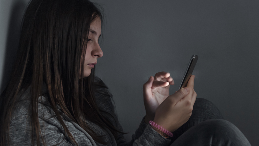 A teenage girl is sitting in a dark room and looking at her cell phone