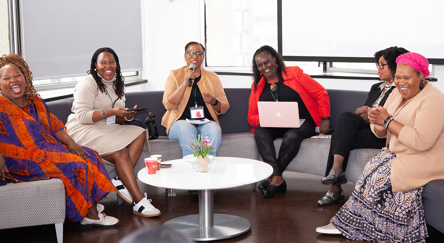 Six Black women are sitting on stage, talking and laughing with big smiles on their face 