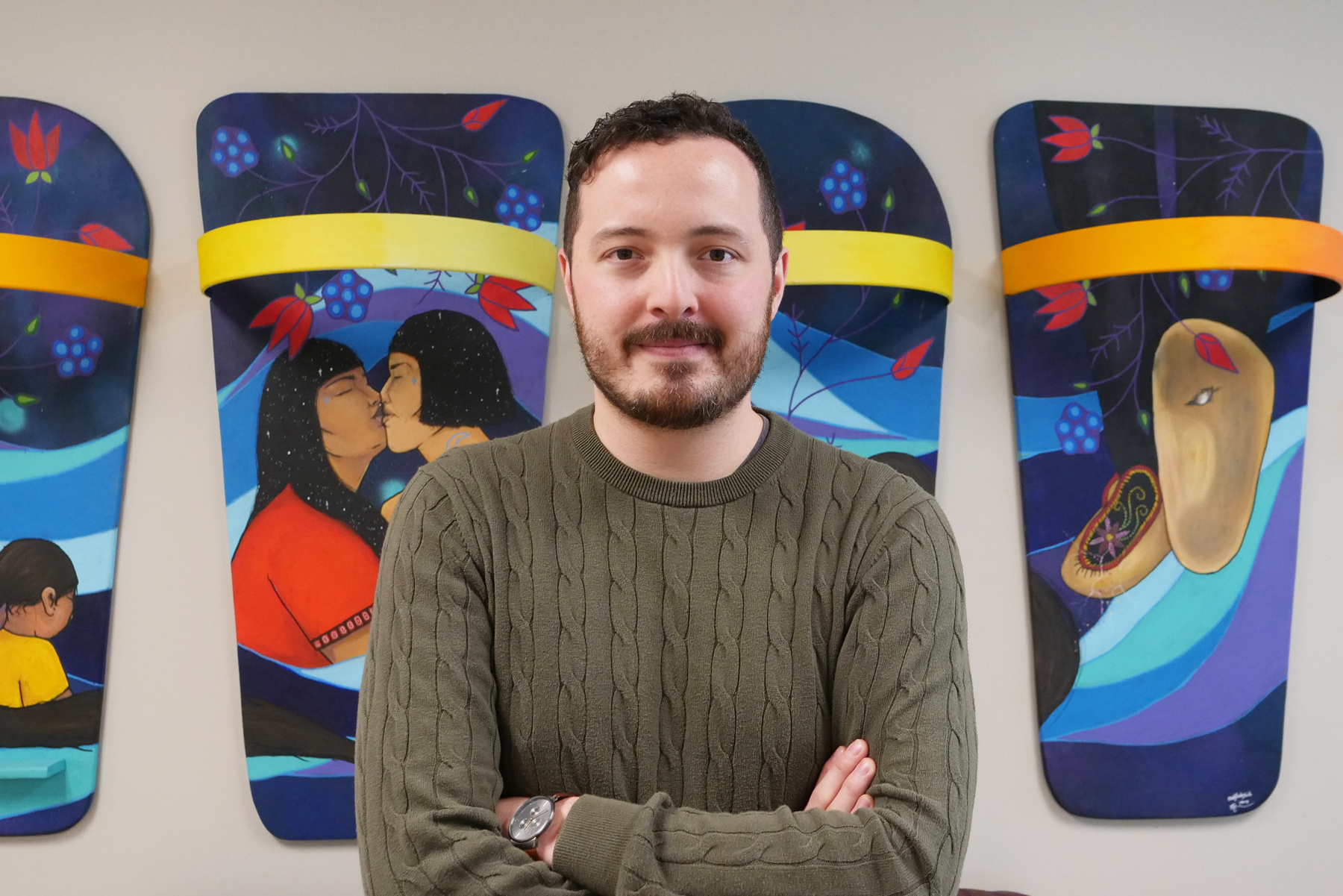 Bearded man standing in green sweater with arms crossed in front of vibrant artwork.