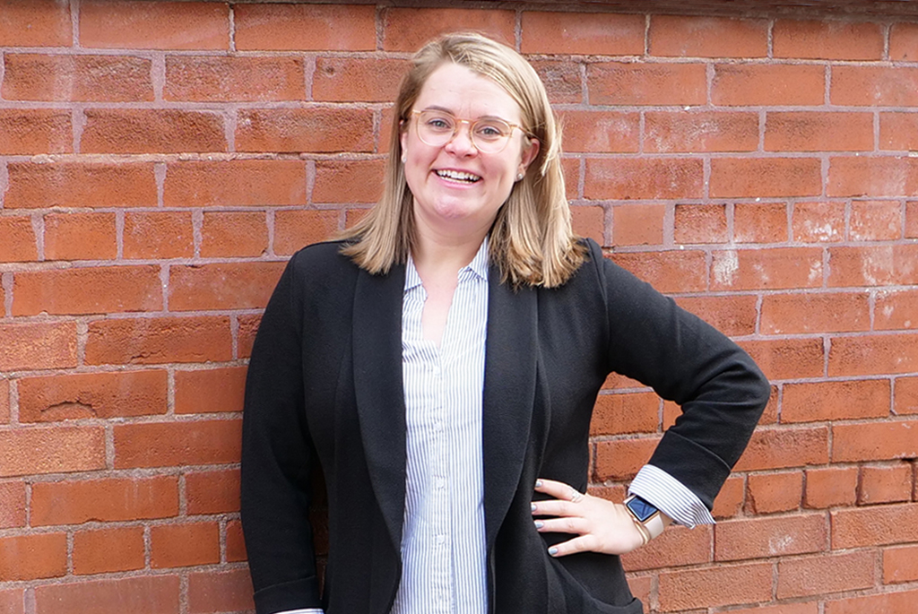 Woman with blond hair smiling with hand on hip in black blazer and blue/white striped shirt.