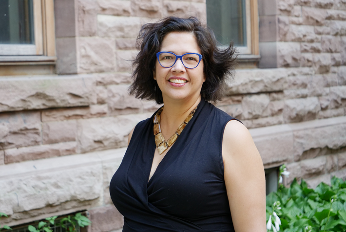 Woman smiling with short hair, blue glasses, black top, and a brown chunky necklace.