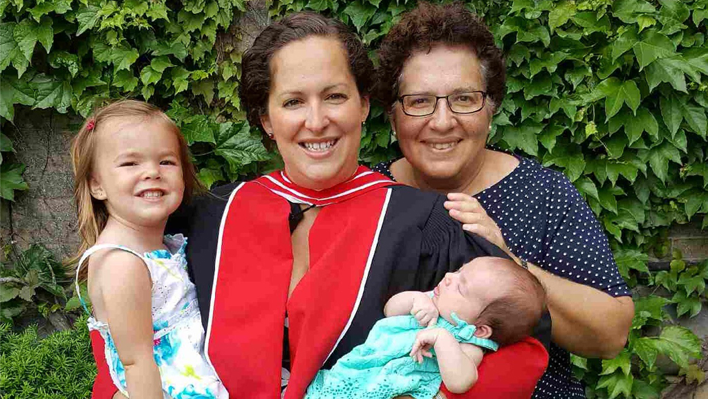 A graduate in a cap and gown smiles while holding a baby, standing with an older woman and a young child.