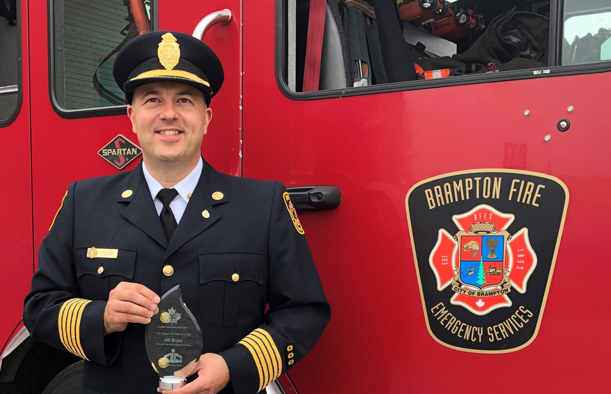 Fire chief in uniform holding an award, standing in front of a Brampton Fire truck.