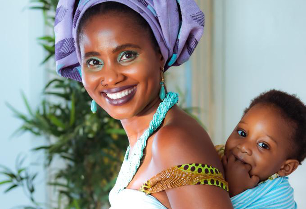 Woman in a purple head wrap smiles while carrying a baby on her back, with green plants in the background.