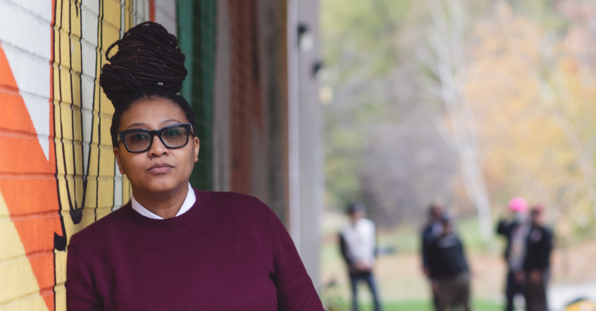  leans against a colorful mural, wearing glasses and a maroon sweater, with an outdoor background.