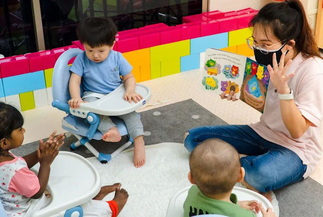 An educator showing a children's book to three babies in highchairs. 