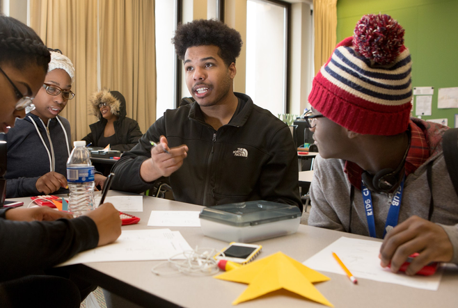 A group of three Black youth are sitting around a table in classroom setting and engaged in converation