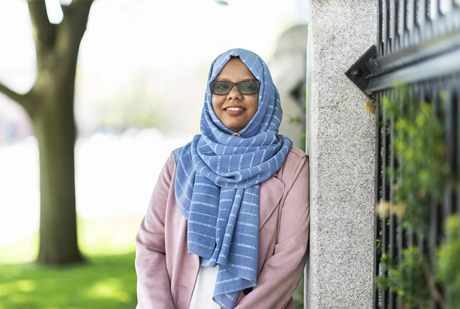 Mariam Abeid is wearing a blue hijab and sunglasses. She is smiling and is casually leaning against a pillar.