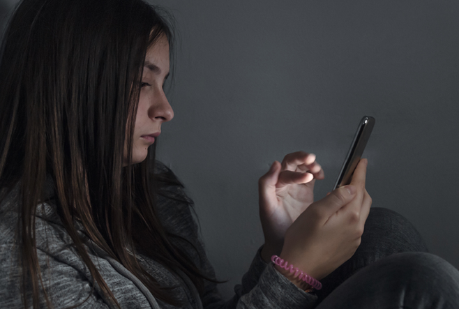 A teenage girl is sitting in a dark room and looking at her cell phone