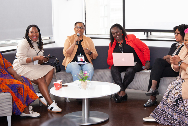 Six Black women are sitting on stage, talking and laughing with big smiles on their face