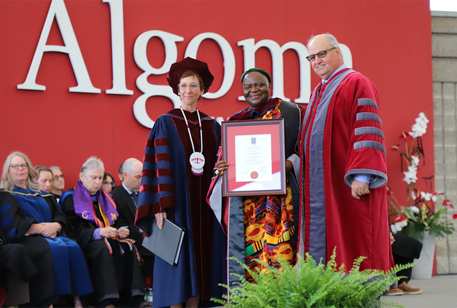 Dr. George Sefa Dei is being honored with an award on stage by two officials from the University of Algoma in academic regalia. They are standing in front of a red backdrop with "Algoma" written on it, with a seated audience behind them.