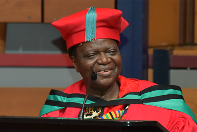 Dr. George Sefa Dei wearing a red academic cap and gown speaks at a podium.