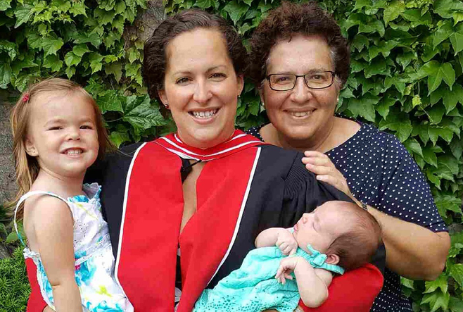 A graduate in a cap and gown smiles while holding a baby, standing with an older woman and a young child.