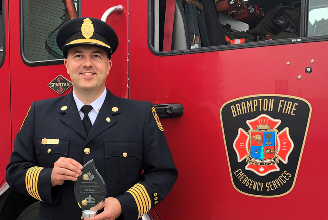 Fire chief in uniform holding an award, standing in front of a Brampton Fire truck.