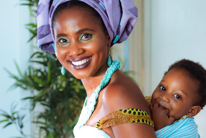 Woman in a purple head wrap smiles while carrying a baby on her back, with green plants in the background.