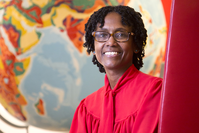  a smiling person in a red outfit with short curly hair and glasses, set against a colorful, stylized globe background.