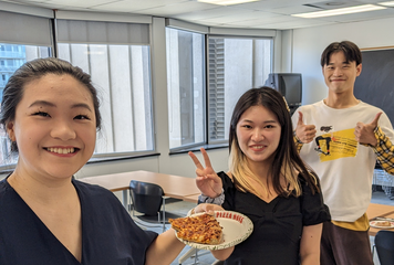 Three people are smiling, one holds pizza, one is giving two thumbs up, and one is making a peace sign.