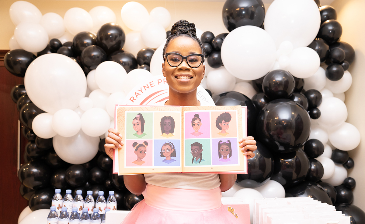 Stephanie Duff, a Black woman, is wearing glasses, proudly holding the book that she authored, and smiling at the camera.