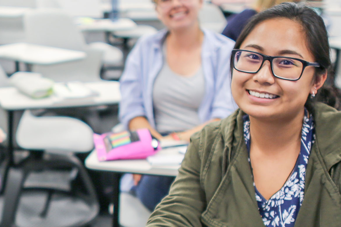 Woman in a green jacket and black glasses sits in a classroom smiling at the camera.