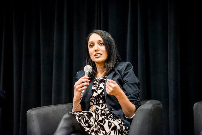 A South Asian woman sitting on a stage and speaking into a microphone