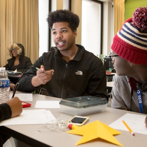 A group of three Black youth are sitting around a table in classroom setting and engaged in converation