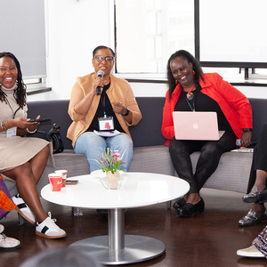 Six Black women are sitting on stage, talking and laughing with big smiles on their face 