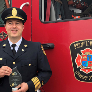 Fire chief in uniform holding an award, standing in front of a Brampton Fire truck.