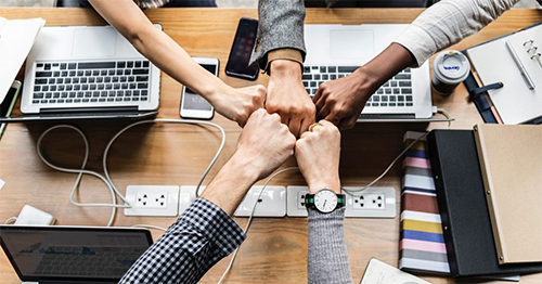 Six touching fists over a desk with their laptops.