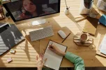 A persons workspace on a wooden table. On the table is a computer, keyboard, notebook, coffee cup and more.