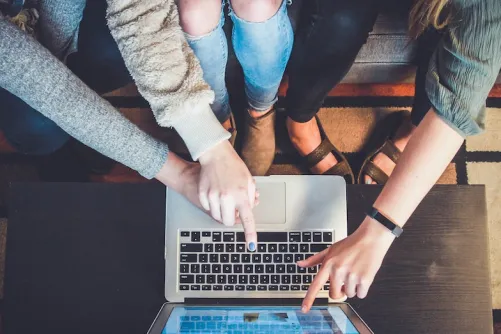 Three people sitting by a laptop. Each of them has one hand on the laptop.