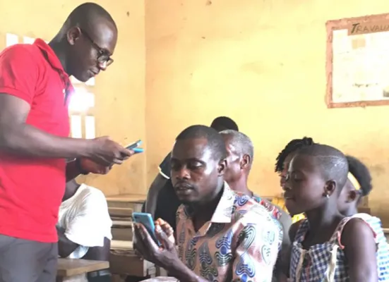 Fabrice Tanoh showing a parent how to use the Allô Alphabet Ed-Tech platform on a mobile phone while his daughter looks on.