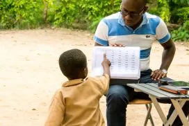 A man seated and holding a paper. A child has their back turned and is pointing at the paper.