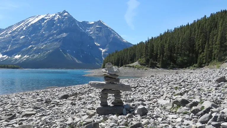 An inuksuk by a body of water, in front of a mountain range.